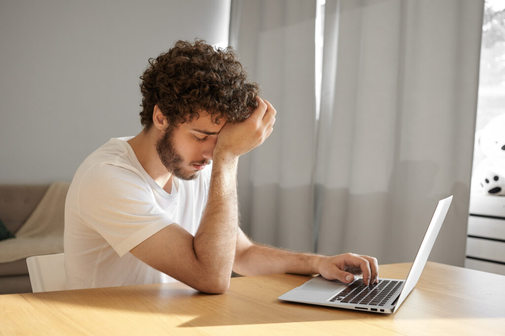 ung fyr (studerende) med brunt krøllet hår og skæg sidder ved et bord og holder sig for panden på grund af hovedpine mens han sidder foran en computer. 