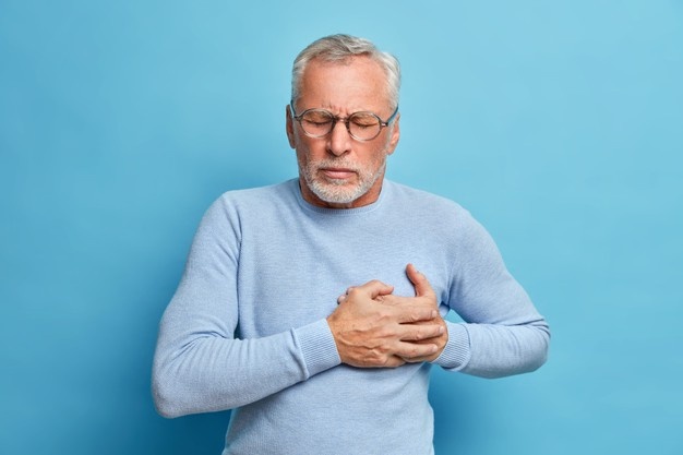 en ældre mand med hvidt hår, briller, lyseblå sweater og hvidt skæg står foran en blå baggrund og holder sig for hjertet i smerte mens han lukker øjnene. 